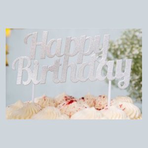 Silver happy birthday cake topper with elegant script lettering displayed on vanilla frosted birthday cake with colorful sprinkles