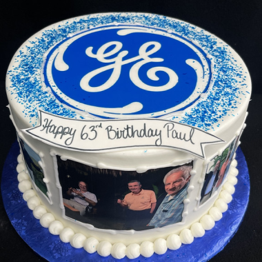 Round edible image cake featuring the iconic GE (General Electric) logo on top with ornate blue mandala border design on edible print. This custom photo cake from Mia's Bakery displays "Happy 65th Birthday Paul" text and black and white portrait photos arranged around the white frosted sides. The corporate celebration cake combines company branding with personal photo memories, finished with blue accents and white piped borders on a blue base.