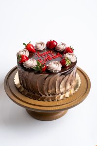7 Layer Birthday Cake from Mia's Bakery featuring rich chocolate layers with glossy chocolate drip frosting on the sides. The birthday cake is topped with alternating swirls of chocolate and vanilla buttercream rosettes, fresh glazed strawberries, and a red 'Happy Birthday!' message written in cursive on the chocolate surface. Displayed on a golden pedestal cake stand against a white background.