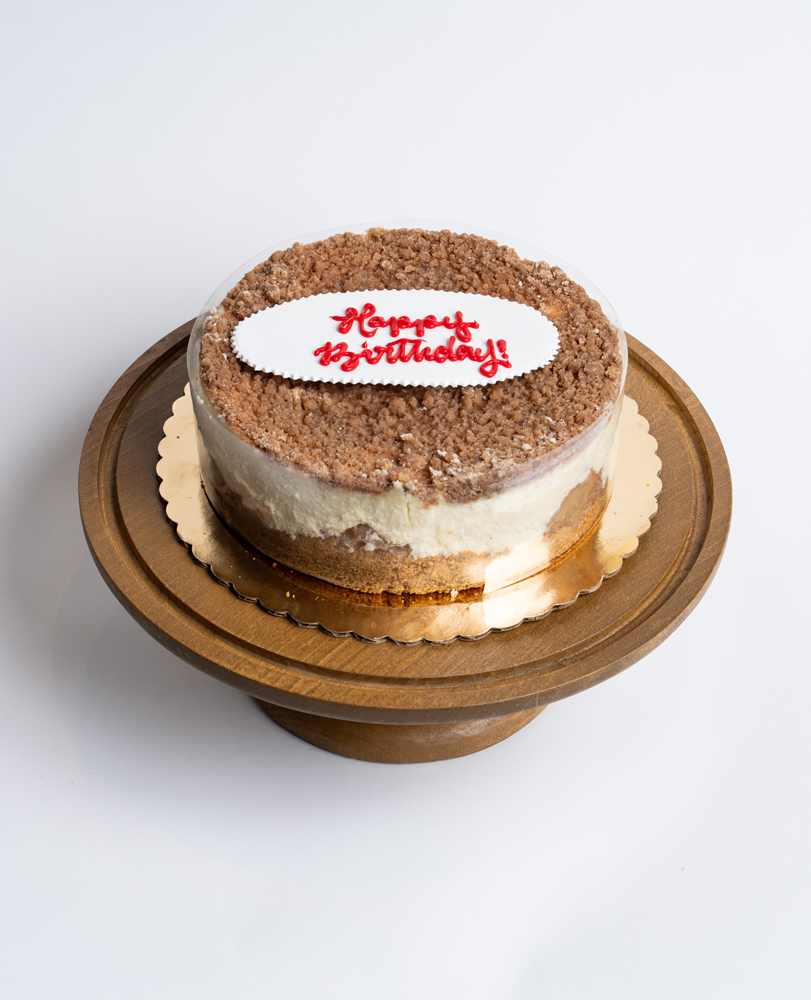 Mia's Bakery Apple Cheesecake Birthday Cake Apple Cheesecake Birthday Cake from Mia's Bakery featuring a round layered cake with creamy white cheesecake filling, golden cake base, and brown cinnamon apple crumble topping. Decorated with a white fondant plaque reading 'Happy Birthday!' in red cursive lettering. The birthday cake is presented on a wooden pedestal cake stand against a white background.