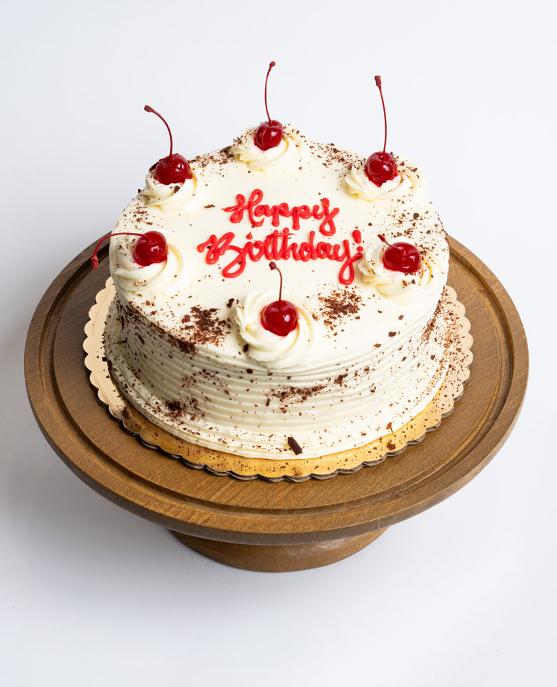 Mia's Bakery Black Forest Birthday Cake Black Forest Birthday Cake from Mia's Bakery featuring a round chocolate layer cake with white whipped cream frosting and horizontal ridged texture. The birthday cake is decorated with piped whipped cream rosettes topped with maraschino cherries with stems around the top edge. Chocolate shavings are dusted over the entire surface. 'Happy Birthday!' is written in red cursive frosting in the center. Presented on a wooden pedestal cake stand against a white background.