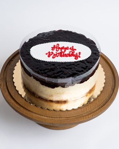 Blueberry Cheesecake Birthday Cake from Mia's Bakery featuring a round cheesecake with creamy white sides and glossy dark blueberry glaze topping. The birthday cake has a deep purple-blue blueberry glaze with visible berry texture covering the top surface. 'Happy Birthday!' is written in red cursive on a white fondant plaque centered on the blueberry topping. The cake has a clear acetate collar around the edge. Displayed on a wooden pedestal cake stand against a white background.