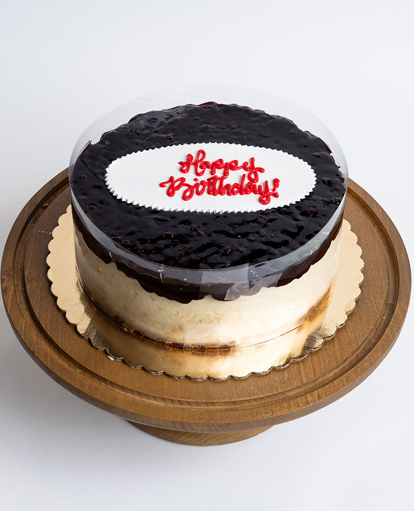Mia's Bakery Blueberry Cheesecake Birthday Cake Blueberry Cheesecake Birthday Cake from Mia's Bakery featuring a round cheesecake with creamy white sides and glossy dark blueberry glaze topping. The birthday cake has a deep purple-blue blueberry glaze with visible berry texture covering the top surface. 'Happy Birthday!' is written in red cursive on a white fondant plaque centered on the blueberry topping. The cake has a clear acetate collar around the edge. Displayed on a wooden pedestal cake stand against a white background.