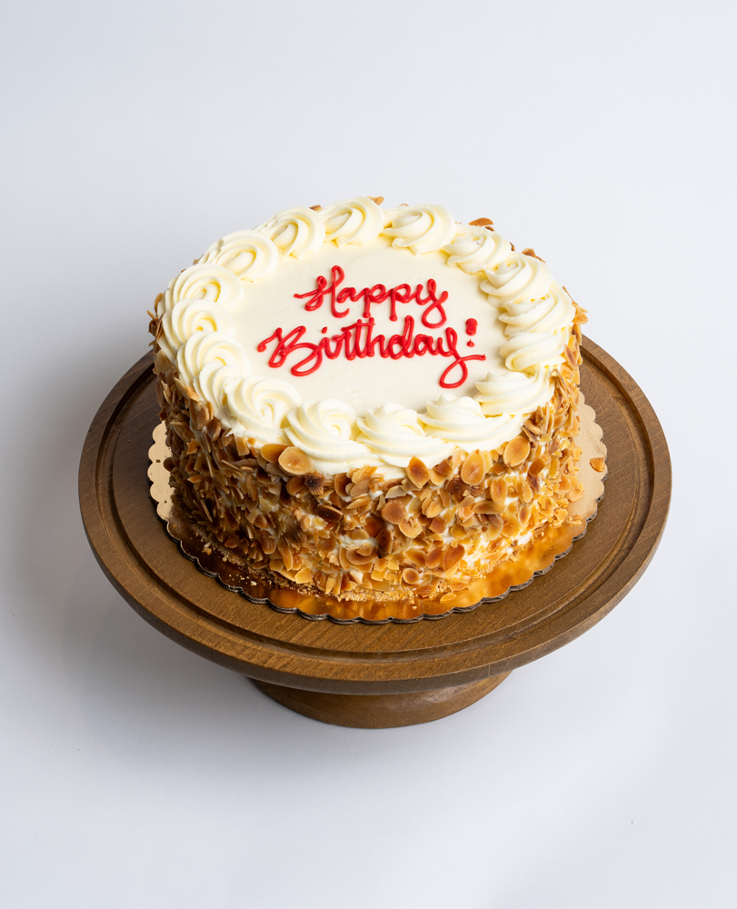 Mia's Bakery Burnt Almond Birthday Cake Burnt Almond Birthday Cake from Mia's Bakery featuring a round layer cake with white buttercream frosting and toasted sliced almonds completely covering the sides. The birthday cake is decorated with piped white buttercream rosettes around the top edge. 'Happy Birthday!' is written in red cursive frosting in the center. The cake has a golden caramel drizzle visible at the base. Presented on a wooden pedestal cake stand against a white background.