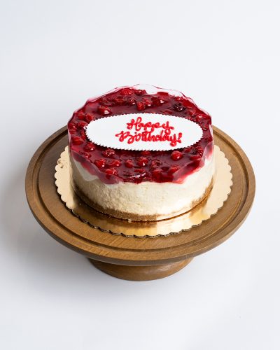 Cherry Cheesecake Birthday Cake from Mia's Bakery featuring a round cheesecake with creamy white sides and glossy red cherry topping. The birthday cake has a ruby-red cherry glaze with visible cherry pieces covering the top surface. 'Happy Birthday!' is written in red cursive on a white fondant plaque centered on the cherry topping. The cake shows a golden graham cracker crust base. Presented on a wooden pedestal cake stand against a white background.