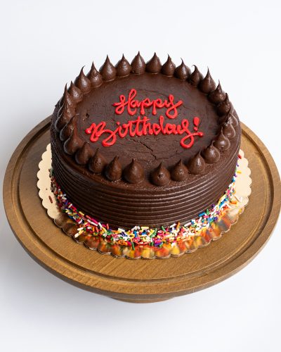 Chocolate Fudge Birthday Cake from Mia's Bakery featuring a round chocolate layer cake completely covered in rich chocolate fudge frosting with horizontal ridged texture on the sides. The birthday cake is decorated with piped chocolate buttercream kisses arranged in a border around the top edge. 'Happy Birthday!' is written in red cursive directly on the chocolate surface. Rainbow sprinkles decorate the bottom edge. Presented on a wooden pedestal cake stand against a white background.