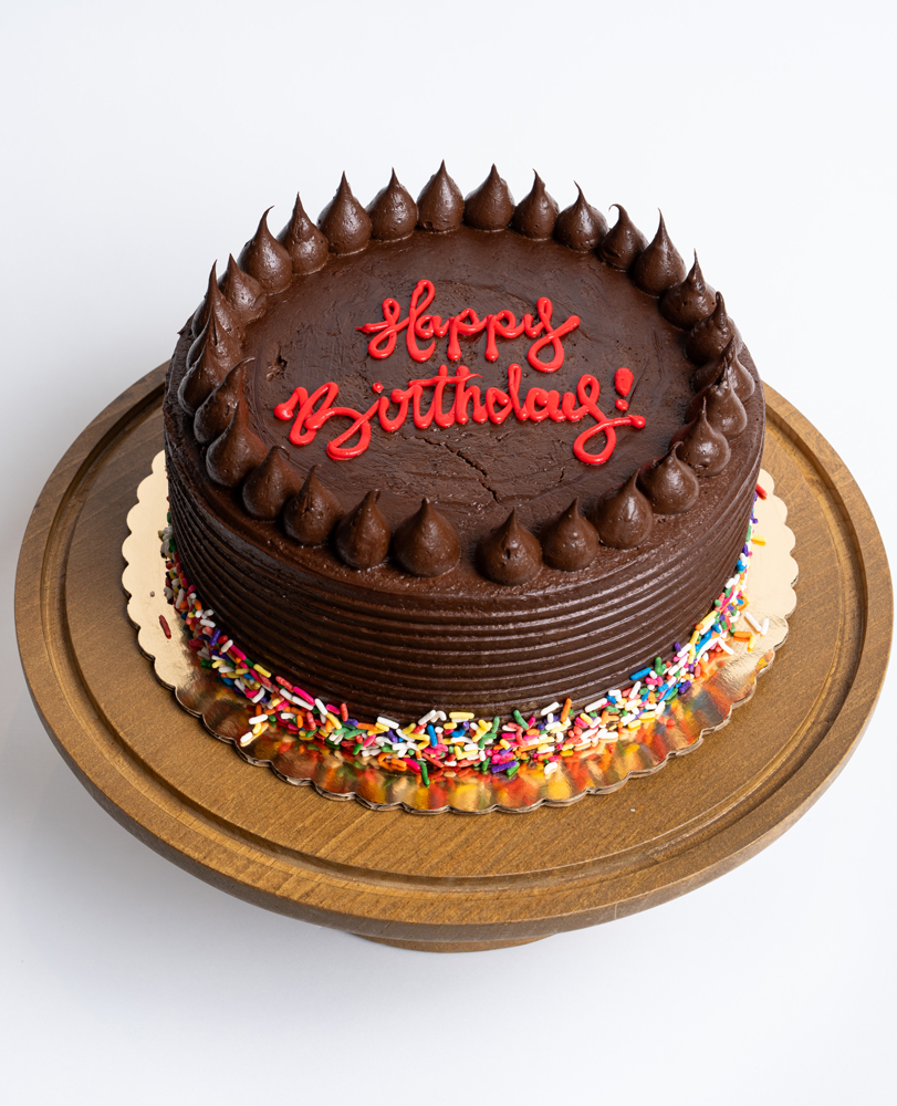 Mia's Bakery Chocolate Fudge Birthday Cake Chocolate Fudge Birthday Cake from Mia's Bakery featuring a round chocolate layer cake completely covered in rich chocolate fudge frosting with horizontal ridged texture on the sides. The birthday cake is decorated with piped chocolate buttercream kisses arranged in a border around the top edge. 'Happy Birthday!' is written in red cursive directly on the chocolate surface. Rainbow sprinkles decorate the bottom edge. Presented on a wooden pedestal cake stand against a white background.