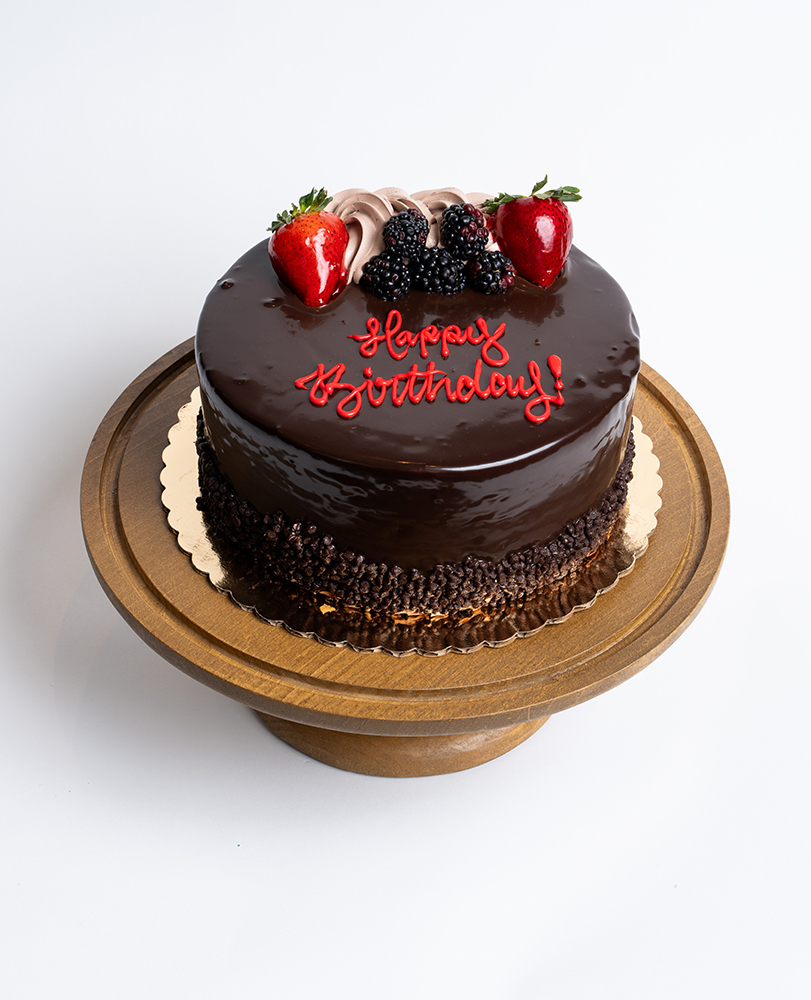 Mia's Bakery Chocolate Layer Birthday Cake Chocolate Layer Birthday Cake from Mia's Bakery featuring a round cake with glossy dark chocolate ganache coating the entire surface. The birthday cake is decorated with fresh strawberries, blackberries, and chocolate buttercream rosettes on top. 'Happy Birthday!' is written in red cursive frosting directly on the chocolate surface. The cake has chocolate pearls or sprinkles around the bottom edge. Displayed on a wooden pedestal cake stand against a white background.