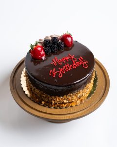 Chocolate Mousse Birthday Cake from Mia's Bakery featuring a round cake with glossy dark chocolate mirror glaze covering the entire top and sides. The birthday cake is decorated with fresh strawberries, blackberries, and chocolate shavings on top, with crushed almonds pressed around the bottom edge. 'Happy Birthday!' is written in red cursive frosting directly on the chocolate surface. Presented on a wooden pedestal cake stand against a white background.