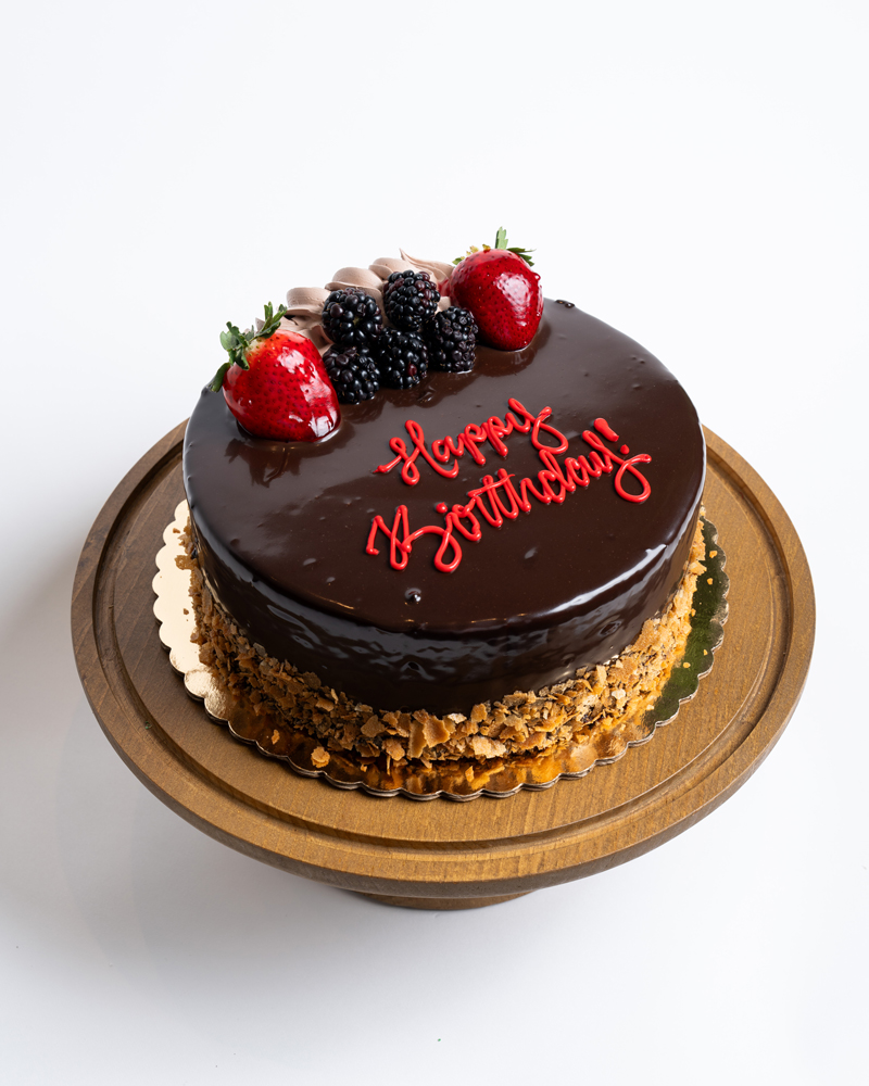 Mia's Bakery Chocolate Mousse Birthday Cake Chocolate Mousse Birthday Cake from Mia's Bakery featuring a round cake with glossy dark chocolate mirror glaze covering the entire top and sides. The birthday cake is decorated with fresh strawberries, blackberries, and chocolate shavings on top, with crushed almonds pressed around the bottom edge. 'Happy Birthday!' is written in red cursive frosting directly on the chocolate surface. Presented on a wooden pedestal cake stand against a white background.