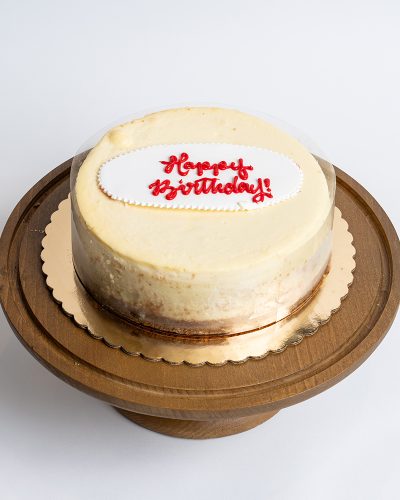 Classic Cheesecake Birthday Cake from Mia's Bakery featuring a round New York style cheesecake with smooth, creamy pale yellow surface and clean sides showing the classic cheesecake texture. The birthday cake has 'Happy Birthday!' written in red cursive frosting on a white fondant plaque placed on top. The cheesecake sits on a golden cake board. Displayed on a wooden pedestal cake stand against a white background.