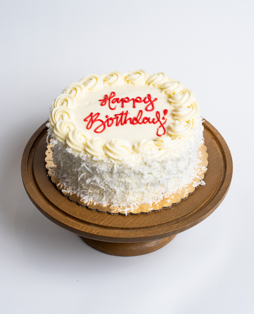 Mia's Bakery Coconut Birthday Cake Coconut Birthday Cake from Mia's Bakery featuring a round single-layer cake completely covered in white buttercream with shredded coconut flakes pressed onto the sides. The birthday cake is decorated with piped vanilla buttercream rosettes around the top edge and displays 'Happy Birthday!' written in red cursive frosting in the center. Presented on a wooden pedestal cake stand against a white background.