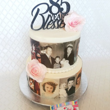 Two-tier white edible image cake featuring vintage black and white family photographs wrapped around both tiers. This custom photo cake from Mia's Bakery displays multiple portraits on edible printed strips, decorated with pink roses and topped with a black "85 Best" cake topper. Perfect for milestone birthdays and anniversary celebrations, this photo cake creates a beautiful memory display using edible image printing technology.