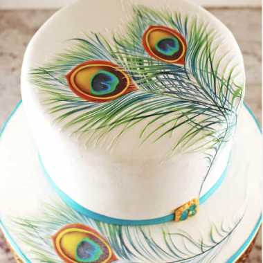 Two-tier white fondant edible image cake featuring vibrant peacock feather designs printed on edible sheets. This artistic photo cake from Mia's Bakery showcases detailed peacock feathers with brilliant blues, greens, and oranges on both tiers, accented with turquoise ribbon trim. The high-quality edible image printing creates a stunning nature-inspired design perfect for elegant celebrations and artistic themed events.