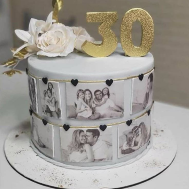 Single-tier round edible image cake featuring a photo collage of black and white couple portraits arranged in a film strip design around the cake sides. This elegant photo cake from Mia's Bakery is decorated with white sugar flowers, gold "50" numbers, and black heart accents. The multiple edible photo prints showcase romantic couple memories, making it perfect for golden anniversary celebrations and milestone birthdays.