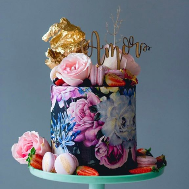 Single-tier artistic edible image cake featuring watercolor-style floral prints on fondant, decorated with pink and purple sugar flowers, gold leaf accents, and fresh berries. This designer photo cake from Mia's Bakery showcases edible printed watercolor roses and peonies wrapped around the cake, topped with macarons and a "Happy 30th" gold topper. A modern take on photo cakes with artistic edible image printing for sophisticated celebrations.