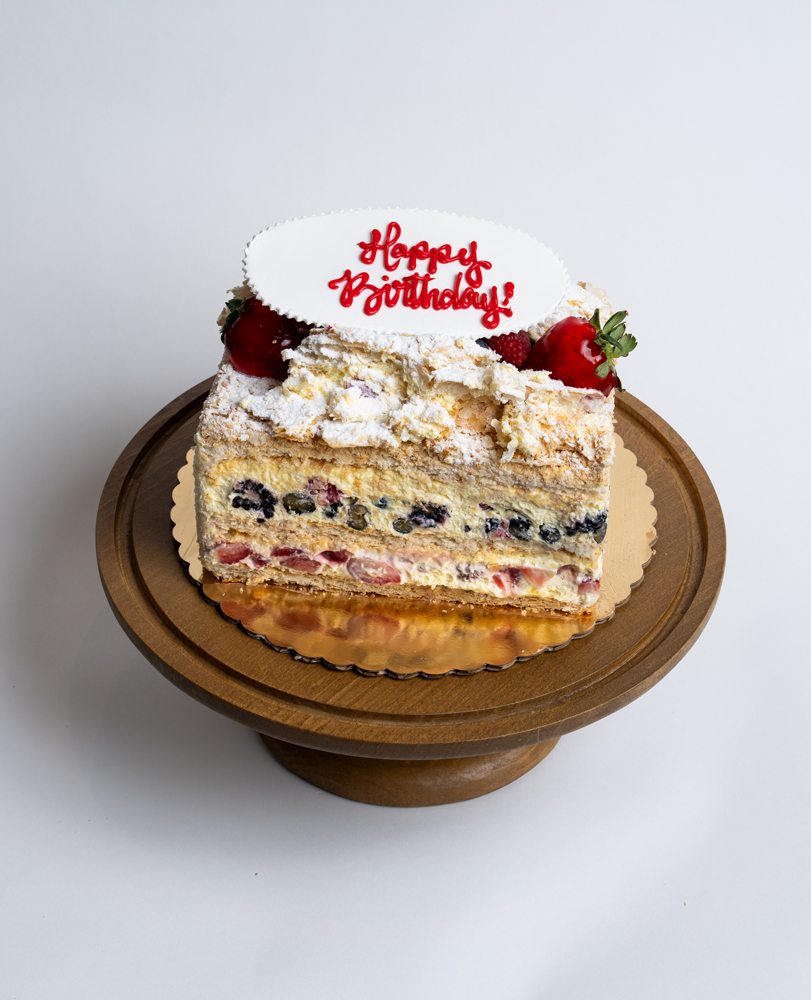 Mia's Bakery Fruit Napoleon Birthday Cake Fruit Napoleon Birthday Cake from Mia's Bakery featuring multiple layers of flaky puff pastry with visible layers containing fresh blueberries, strawberries, and pastry cream filling. The birthday cake is topped with powdered sugar, whipped cream dollops, and fresh strawberries. 'Happy Birthday!' is written in red cursive on a white fondant plaque placed on top. The Napoleon shows golden caramelized base. Presented on a wooden pedestal cake stand against a white background.