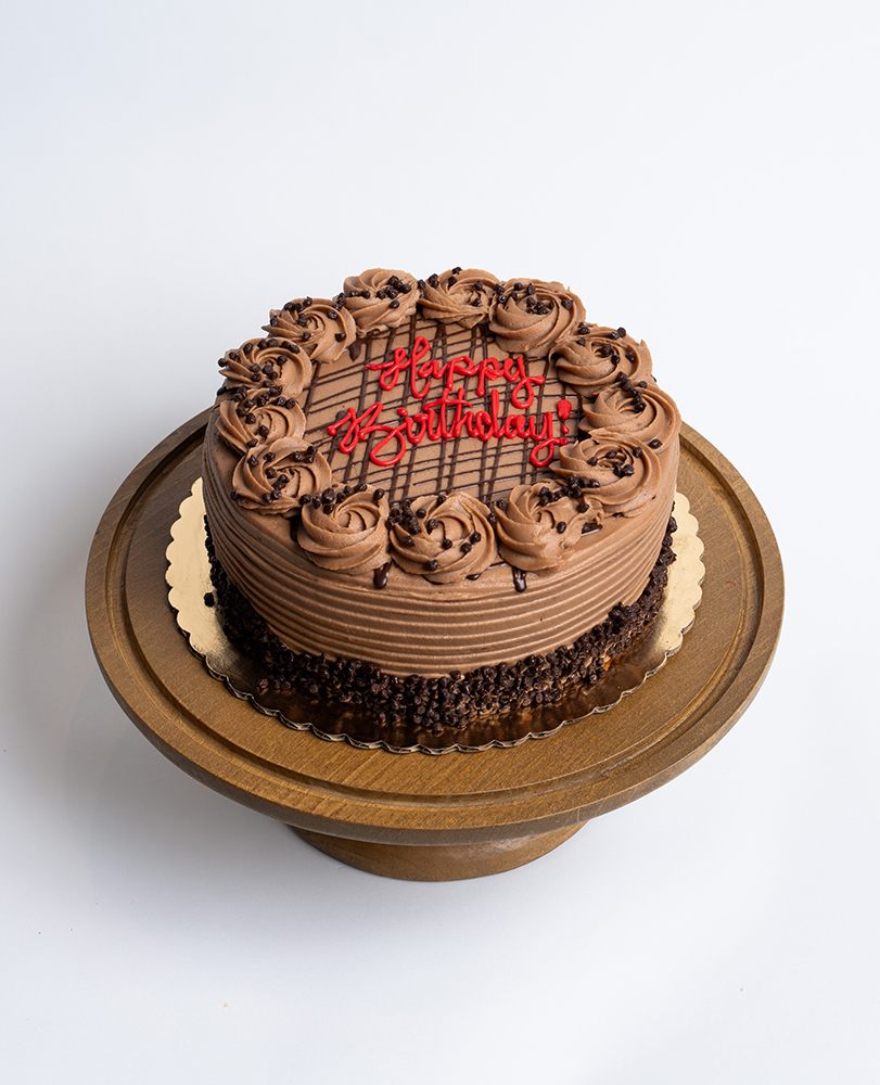 Mia's Bakery Nutella Birthday Cake Nutella Birthday Cake from Mia's Bakery featuring a round chocolate layer cake completely covered in rich Nutella-style chocolate buttercream with horizontal ridged texture on the sides. The birthday cake is decorated with piped chocolate buttercream rosettes around the top edge and has chocolate crumbs pressed around the bottom edge. The top features chocolate drizzle lines and 'Happy Birthday!' written in red cursive frosting. Displayed on a wooden pedestal cake stand against a white background.