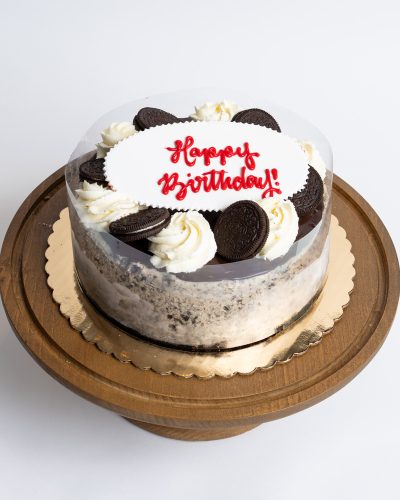 Oreo Cheesecake Birthday Cake from Mia's Bakery featuring a round cheesecake with white cream filling and visible Oreo cookie crumb layers on the sides. The birthday cake is decorated with whole and half Oreo cookies alternating with piped vanilla buttercream rosettes around the top edge. 'Happy Birthday!' is written in red cursive on a white fondant plaque in the center. The cake has a clear acetate collar around the sides. Displayed on a wooden pedestal cake stand against a white background.