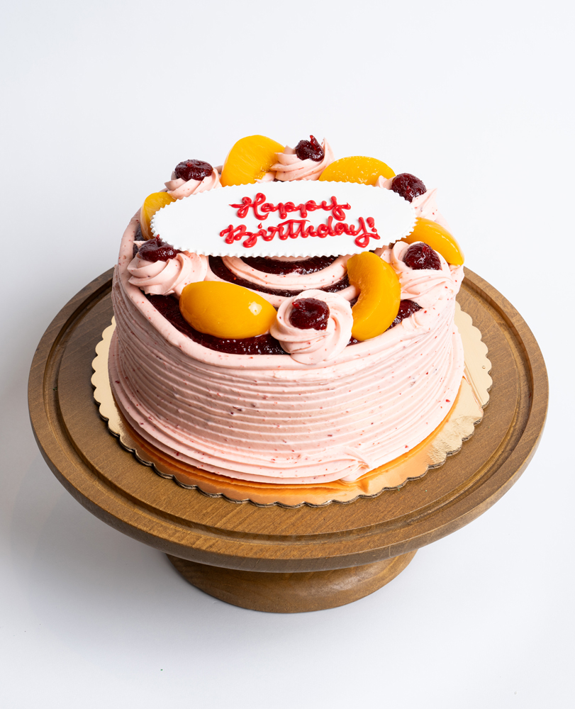 Mia's Bakery Peach Raspberry Birthday Cake Peach Raspberry Birthday Cake from Mia's Bakery featuring a round single-layer cake covered in pink raspberry buttercream with horizontal ridged texture on the sides. The birthday cake is decorated with fresh peach slices, raspberry jam dollops, piped pink buttercream rosettes, and dark chocolate cake layers visible through decorative openings. 'Happy Birthday!' is written in red cursive on a white fondant plaque placed on top. Presented on a wooden pedestal cake stand against a white background.