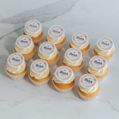 Collection of twelve vanilla edible image cupcakes featuring circular Alainn brand logos printed on white fondant toppers. These custom photo cupcakes from Mia's Bakery have orange and yellow swirled buttercream frosting with professional edible printed logos centered on each cupcake. Displayed on marble surface, these branded cupcakes are perfect for corporate events, product launches, and company celebrations.RetryClaude can make mistakes. Please double-check responses.