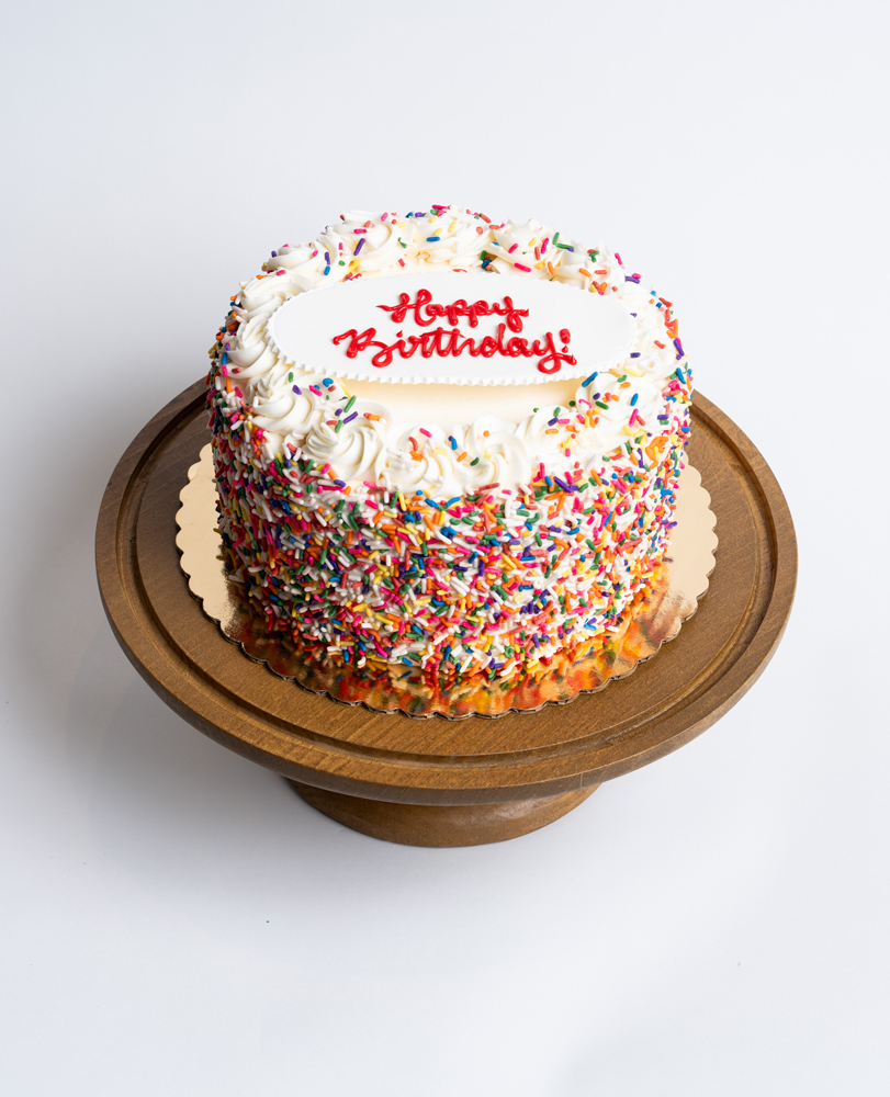 Mia's Bakery Rainbow Birthday Cake Rainbow Birthday Cake from Mia's Bakery featuring a round single-layer cake covered in white vanilla buttercream with multicolored rainbow sprinkles completely coating the sides. The birthday cake is decorated with piped white buttercream rosettes around the top edge, each topped with rainbow sprinkles. 'Happy Birthday!' is written in red cursive frosting on a white fondant plaque centered on top. Displayed on a wooden pedestal cake stand against a white background.