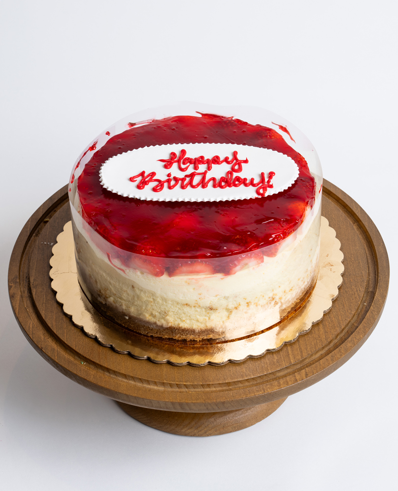 Mia's Bakery Strawberry Cheesecake Birthday Cake Strawberry Cheesecake Birthday Cake from Mia's Bakery featuring a round cheesecake with creamy white sides and glossy red strawberry glaze topping. The birthday cake displays 'Happy Birthday!' written in red cursive on a white fondant plaque centered on the strawberry glaze. The cake shows visible layers with a golden graham cracker crust base. Presented on a wooden pedestal cake stand against a white background.