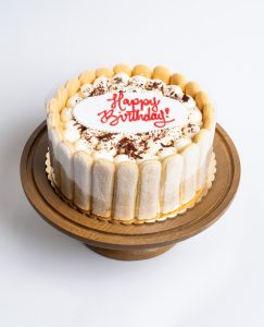 Tiramisu Birthday Cake from Mia's Bakery featuring a round cake decorated with ladyfinger cookies standing vertically around the sides. The birthday cake has a white cream top dusted with cocoa powder and chocolate shavings, with 'Happy Birthday!' written in red cursive frosting in the center on a white fondant plaque. Displayed on a wooden pedestal cake stand against a white background.