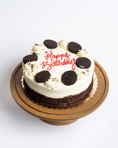 Oreo Birthday Cake from Mia's Bakery featuring a round chocolate layer cake with white vanilla buttercream on top and visible dark chocolate cake sides. The birthday cake is decorated with whole Oreo cookies arranged in a circle around the top edge, interspersed with piped white buttercream swirls and Oreo cookie crumb sprinkles. 'Happy Birthday!' is written in red cursive frosting in the center. Presented on a wooden pedestal cake stand against a white background.