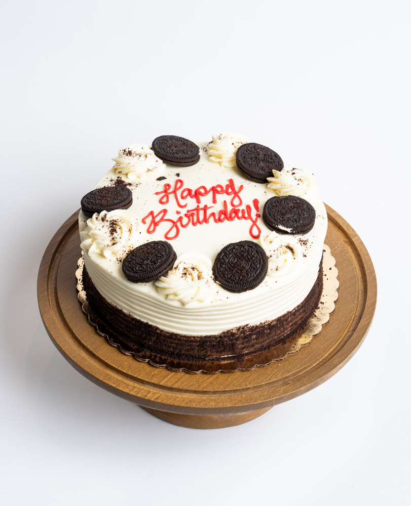 Mia's Bakery Oreo Birthday Cake Oreo Birthday Cake from Mia's Bakery featuring a round chocolate layer cake with white vanilla buttercream on top and visible dark chocolate cake sides. The birthday cake is decorated with whole Oreo cookies arranged in a circle around the top edge, interspersed with piped white buttercream swirls and Oreo cookie crumb sprinkles. 'Happy Birthday!' is written in red cursive frosting in the center. Presented on a wooden pedestal cake stand against a white background.