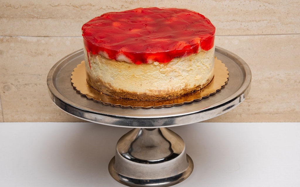 Strawberry cheesecake with glossy red fruit topping on graham cracker crust displayed on silver cake stand