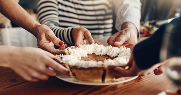 Family hands reaching for thanksgiving desserts including pumpkin pie with whipped cream at holiday gathering on wooden table