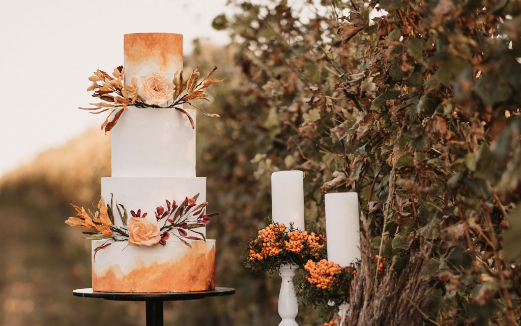 Fall wedding cake with hand-painted orange buttercream, dried flowers, and candles against autumn foliage