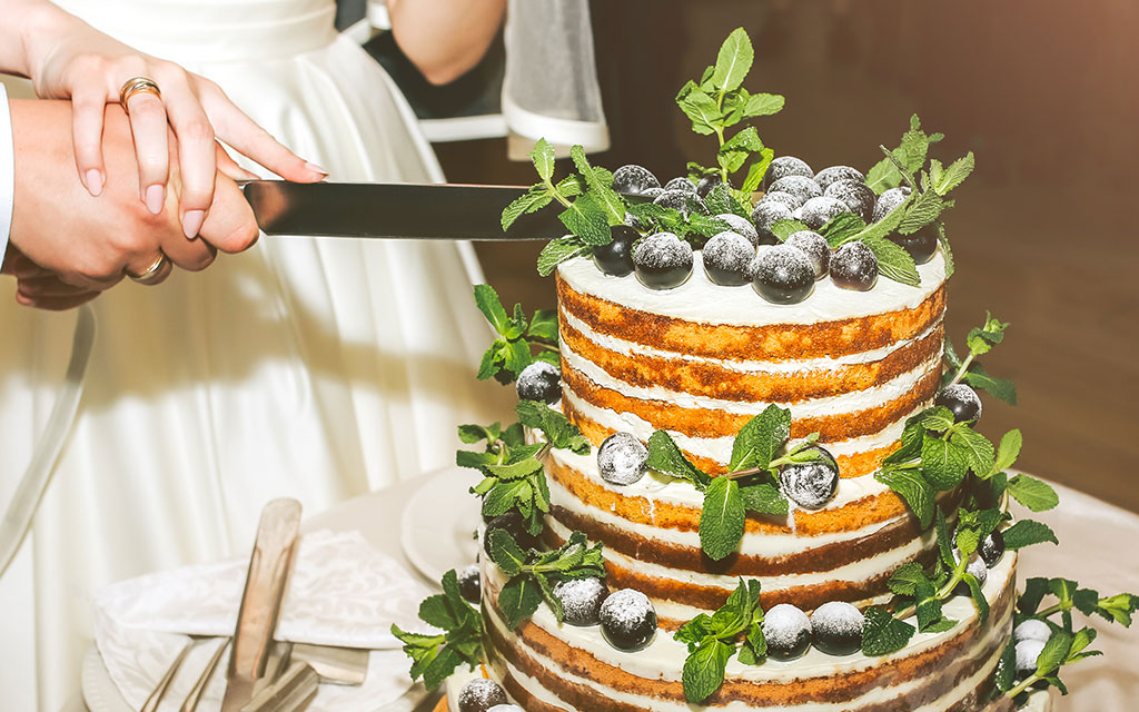 Bride cutting naked fall wedding cake with exposed layers, fresh berries, and mint garnish at reception