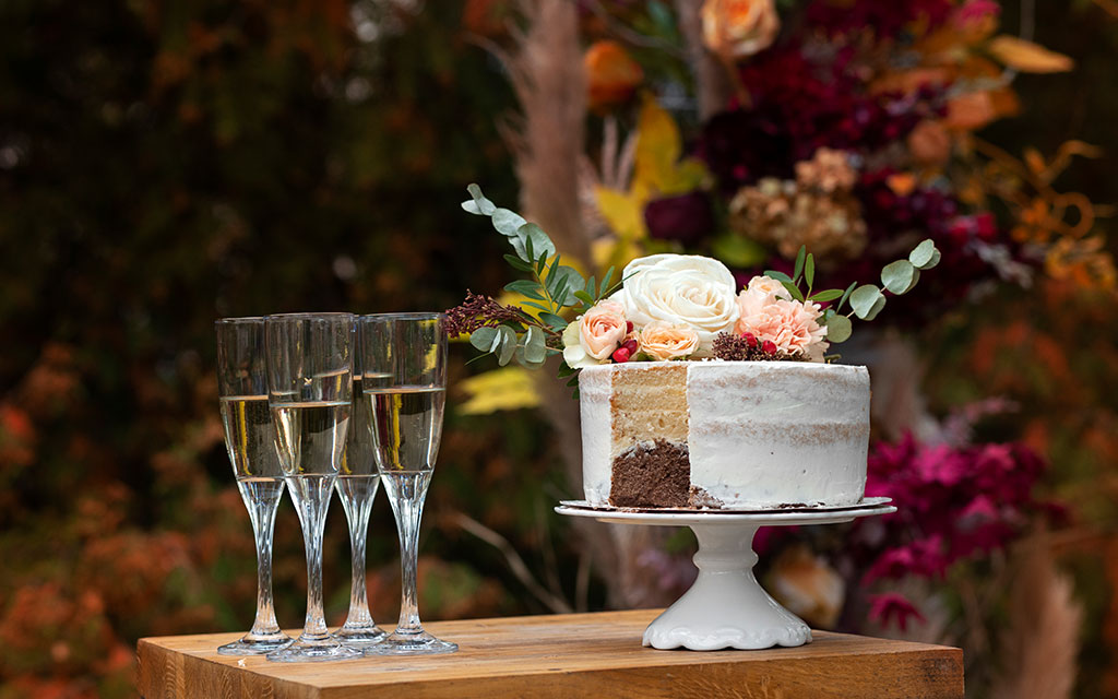 Semi-naked fall wedding cake with fresh roses, eucalyptus, and berries on white stand with champagne